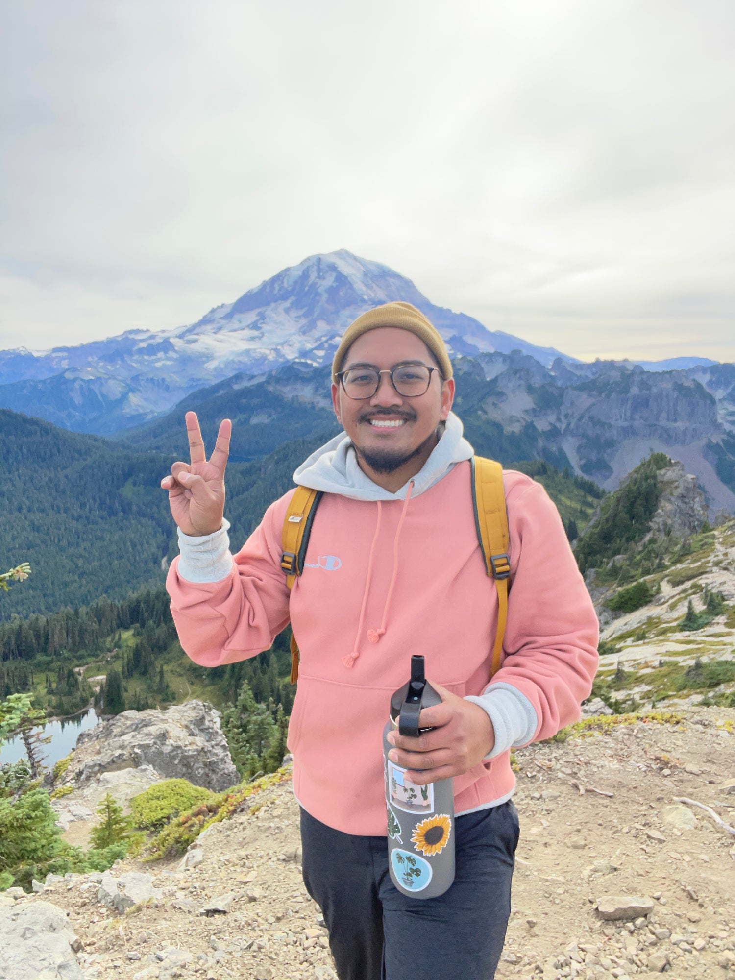 Person in a pink jacket with a yellow strap, holding a bottle, standing in front of a mountainous landscape.